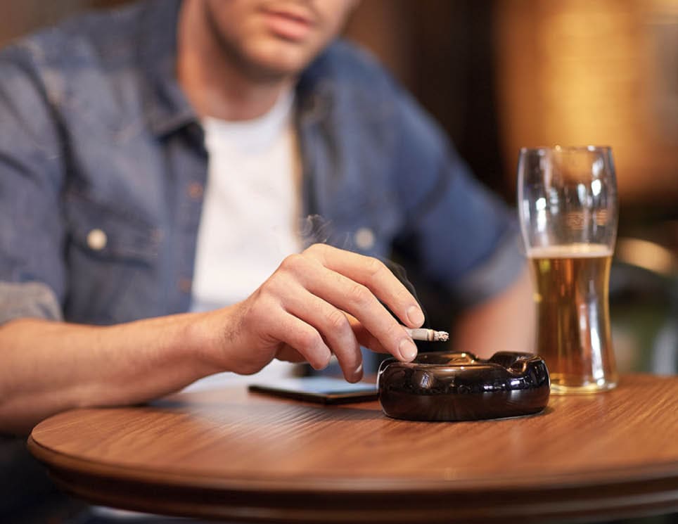 people, nicotine addiction and bad habits concept - close up of man drinking beer, smoking cigarette and shaking ashes to ashtray at bar or pub