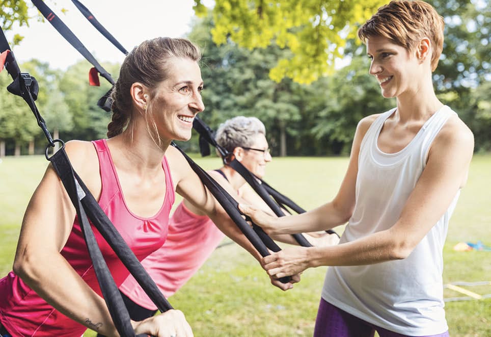 Smiling young woman doing exercises using suspension training system with female trainer in park